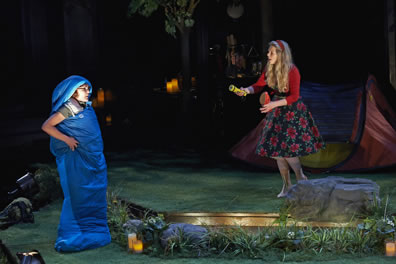 Lysander stands while wrapped up in a blue sleeping bag as she talks to Helena, one step up, wearing a large-roses-print skirt and red sweater with read hair band and carrying a flashlight. The stage has grass with weeds around the steps and a large rock to the right, and red tent with blue screen windows is behind Helena